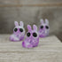 Three small, purple Plasma Storm Chenille figures from Bindlewood Shop, with big black eyes and a glossy, marbled finish, are arranged on wood. Their cute design is inspired by Amanda Louise Spayd and evokes a whimsical charm.