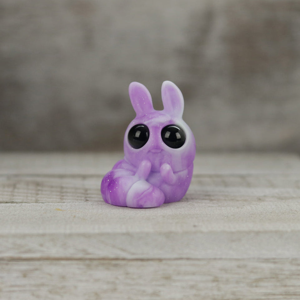 Plasma Storm Chenille, a small purple bunny-like resin figure by Bindlewood Shop and Amanda Louise Spayd, sits on a wooden surface with large black eyes and raised front paws. The background is blurred light brown wood.