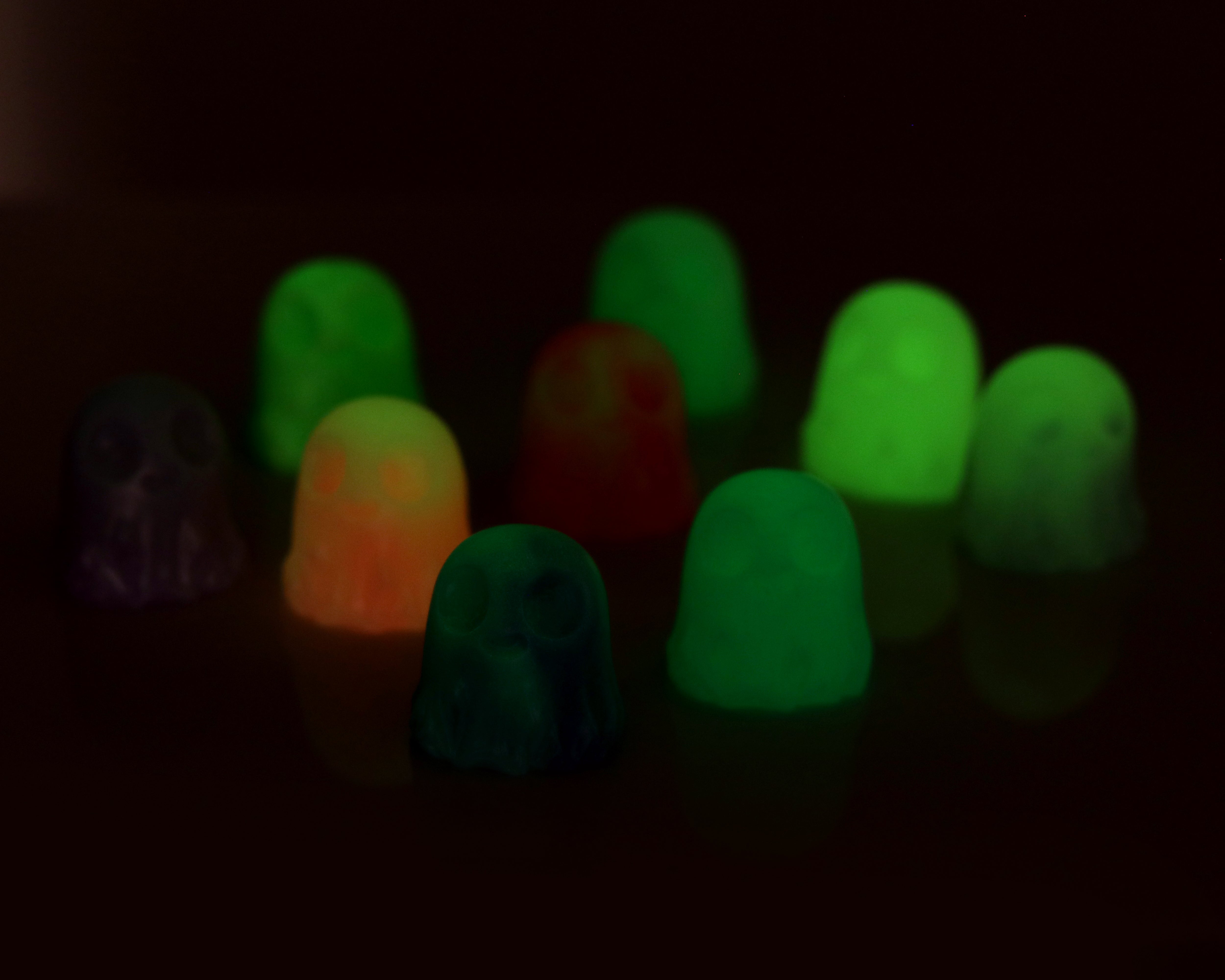 Two Giddy Ghost Glow figurines by Bindlewood Shop, designed by Chris Ryniak, emit a glowing light in the dark and feature shades of green, orange, red, and purple. They are displayed together against a dark background.