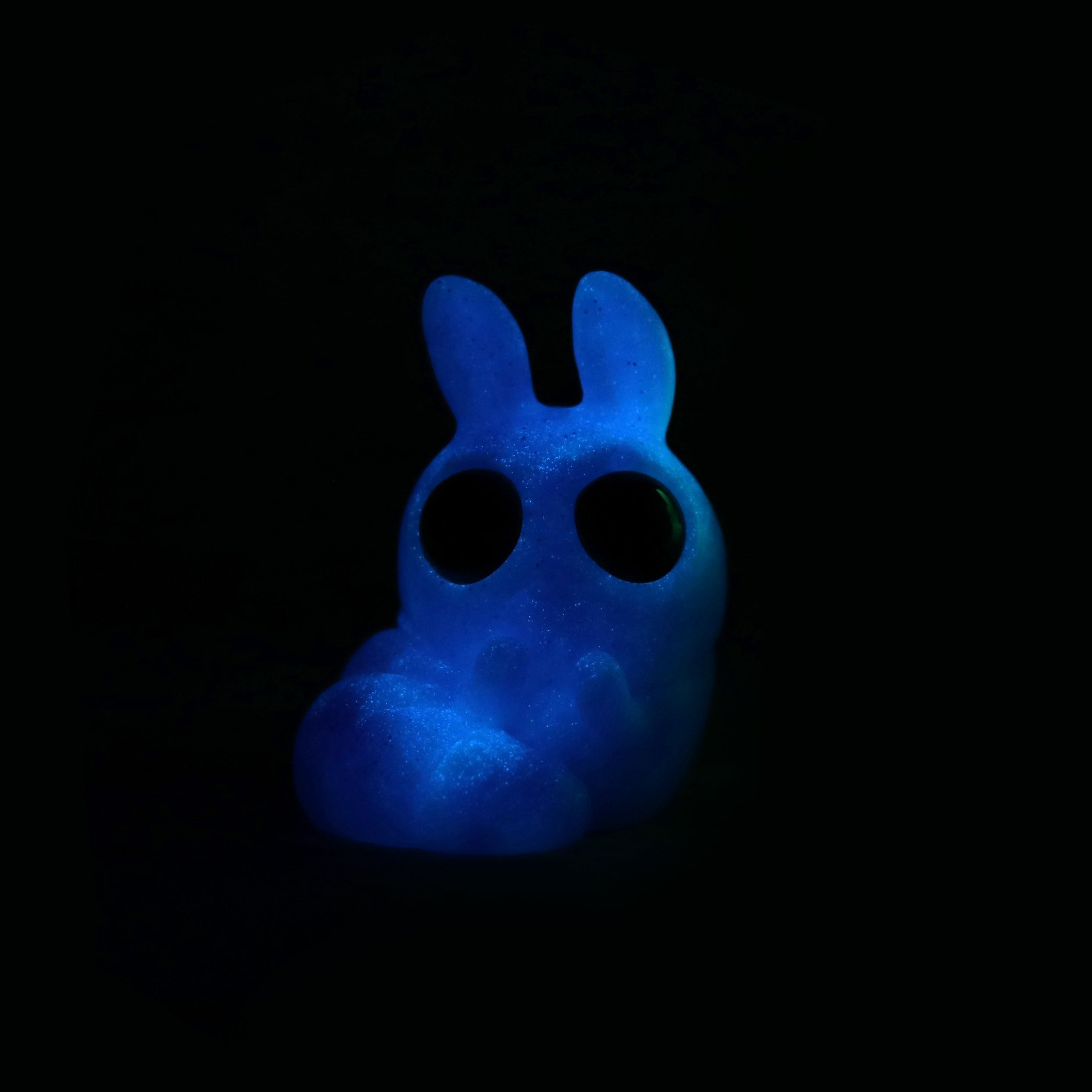 Plasma Storm Chenille by Bindlewood Shop is a small, bunny-shaped resin figure with large round eyes that glows blue in the dark, set against a black background, inspired by Amanda Louise Spayd’s whimsical style.
