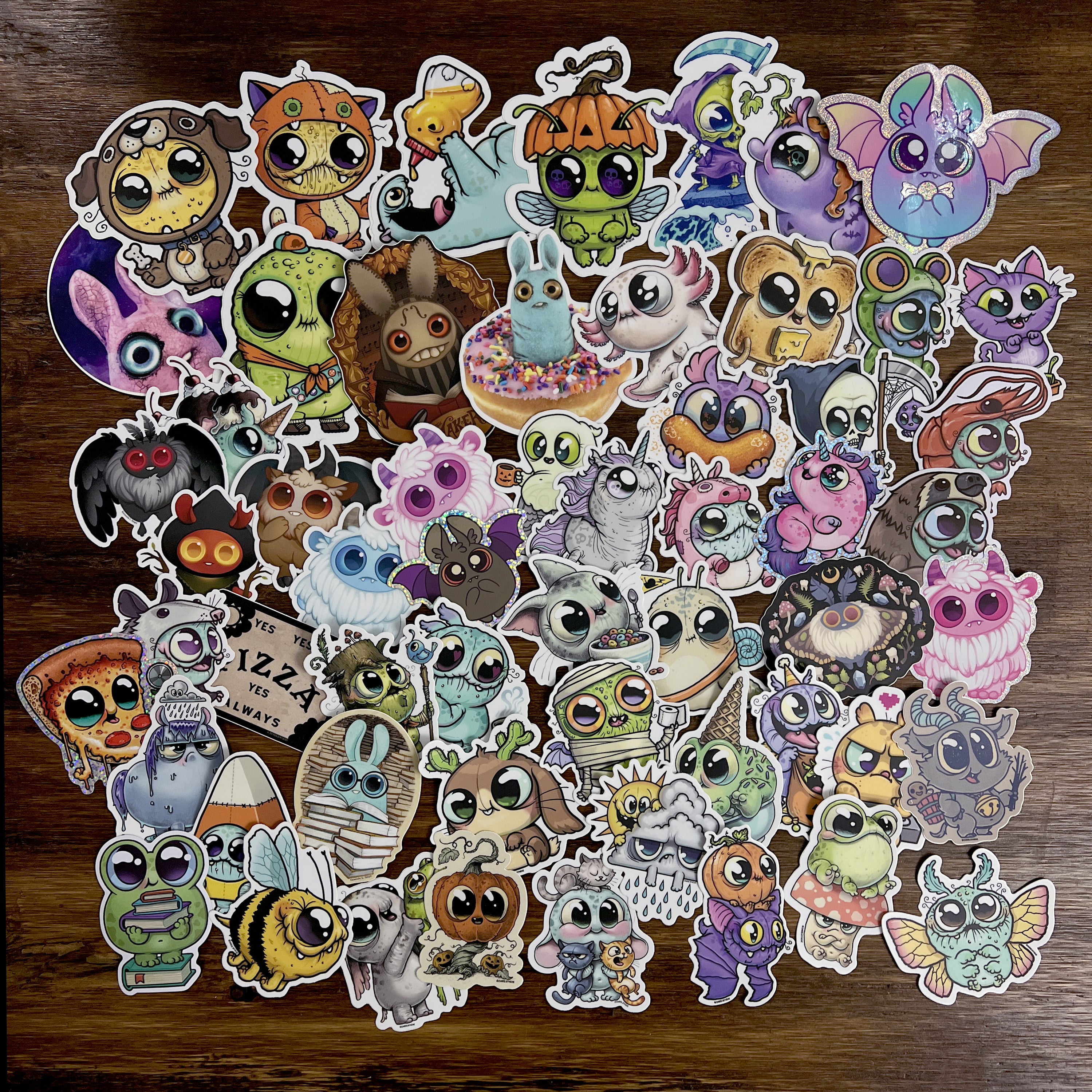 A vibrant ULTRA MEGA STICKER PACK! from Bindlewood Shop is displayed on a wooden surface, showcasing cartoon-style animal stickers with large, expressive eyes dressed in whimsical costumes like mummies, pumpkins, and bees. This playful collection is perfect for adding charm to your favorite items.