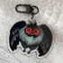 The Bindlewood Shop Mothman Keychain features a double-sided acrylic charm depicting a cartoon bat-like creature with big red eyes, fluffy grey fur, and black wings. Presented on a light wood surface, this keychain includes a coated metal clip for easy attachment.