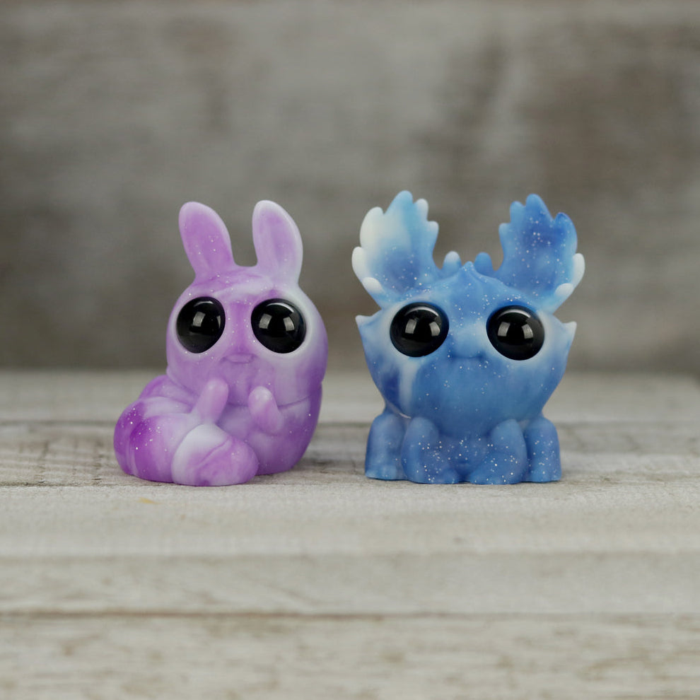 The Bindlewood Shop Sunshower Moose & Plasma Storm Chenille Set features two small, glittery resin figures with large black eyes—a purple bunny and a Caribbean Blue Dashi-inspired moose—side by side on a wooden surface.