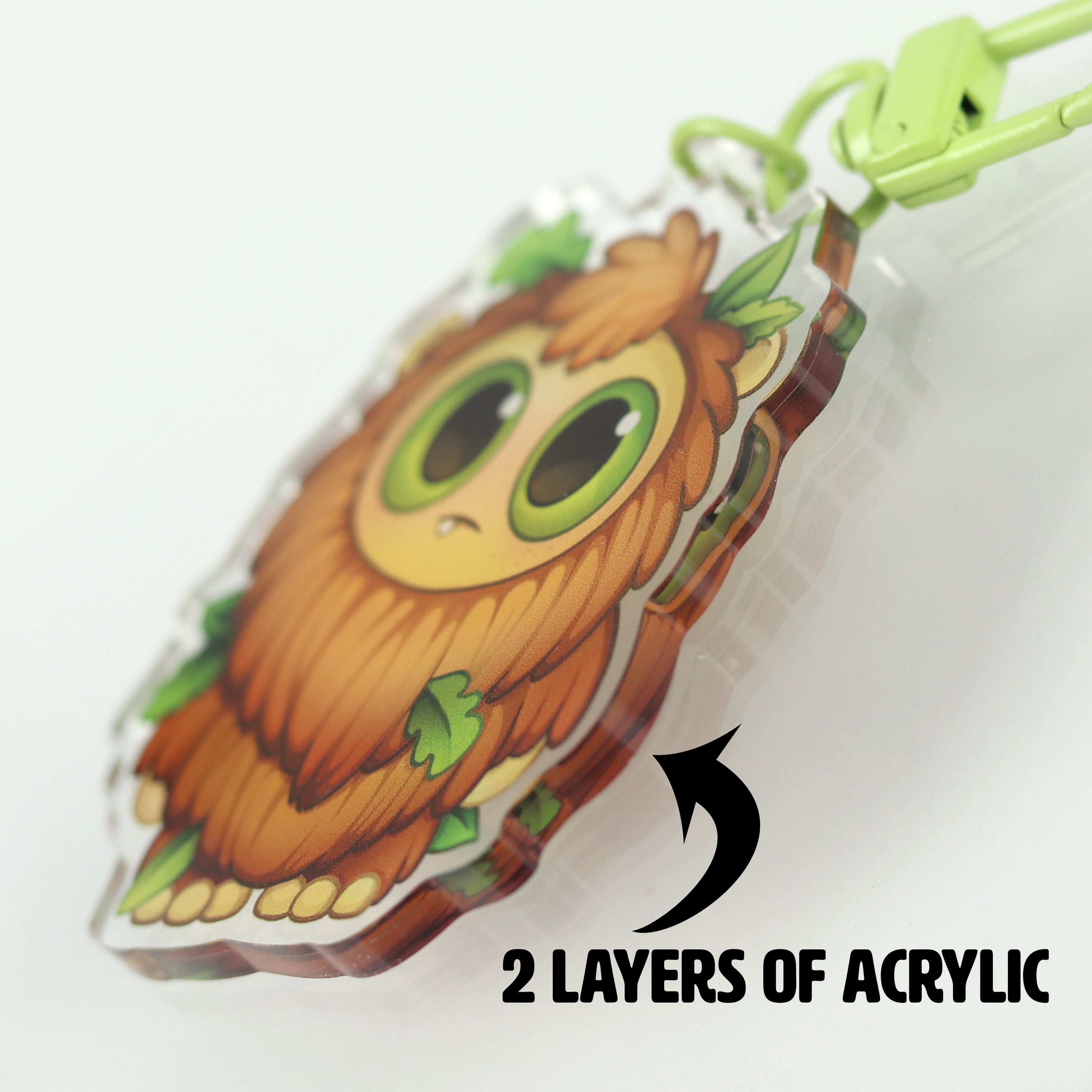 A close-up of "The Maker" keychain by Bindlewood Shop, owl-shaped with two acrylic layers, large green eyes, orange-brown feathers with green leaves, green clasp. Text reads “2 LAYERS OF ACRYLIC.”.
