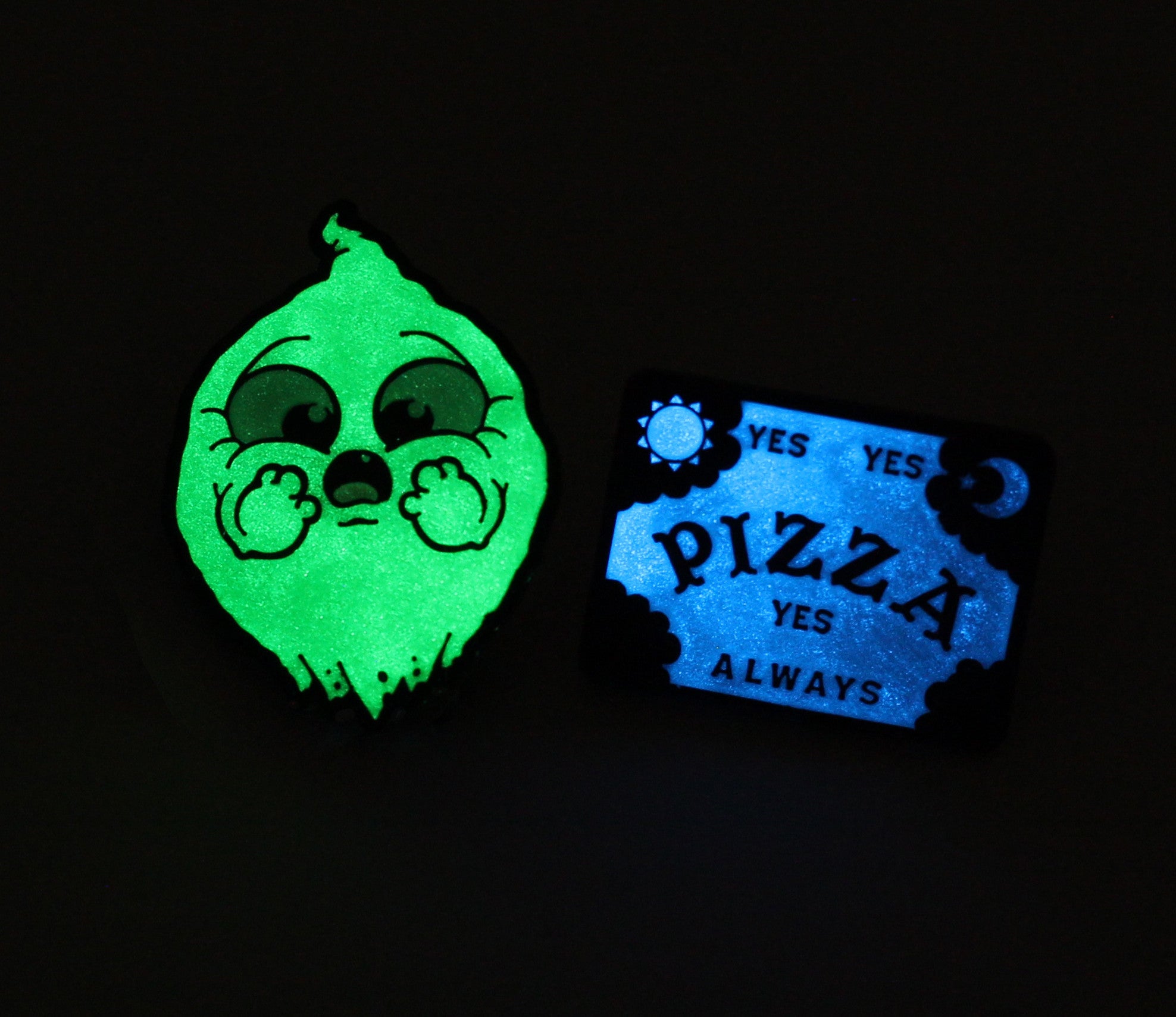 The OMGhost Pin Set by Bindlewood Shop features a glow-in-the-dark green cartoon ghost with chubby cheeks holding a blue spirit board pin that says “YES PIZZA YES ALWAYS,” accented with sun and moon details on a black background.