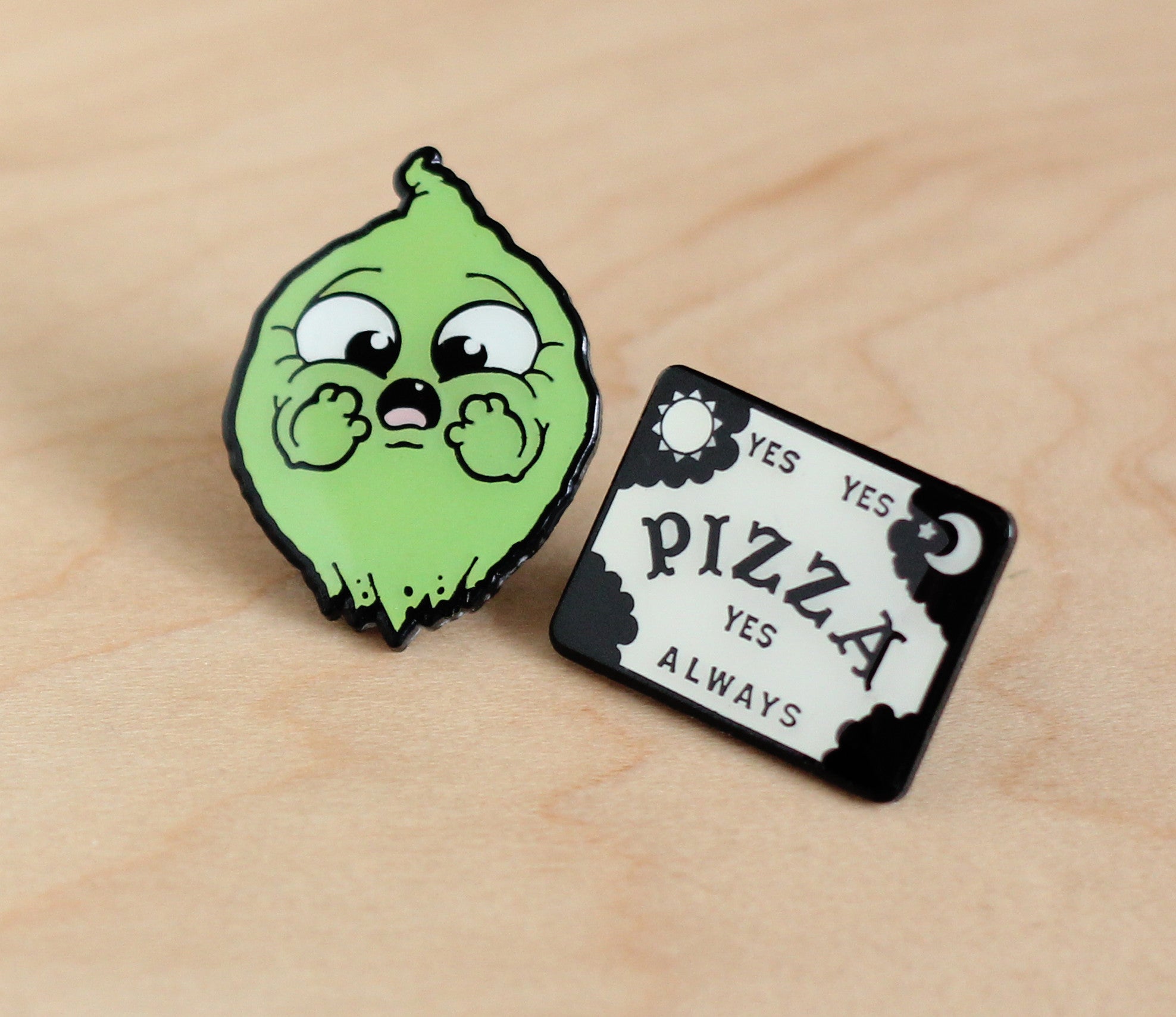 The Bindlewood Shop OMGhost Pin Set features two enamel pins on wood: a cute, wide-eyed green lemon with tiny hands on its cheeks, and a glow-in-the-dark pin reading "YES YES PIZZA YES ALWAYS" in bold black letters.