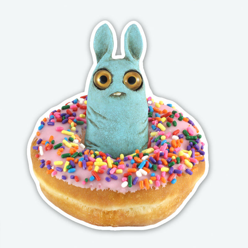 A blue, wide-eyed Pipsqueak with bunny ears sits in a pink-frosted, sprinkle-topped donut in the Donut Pip Sticker by Bindlewood Shop.