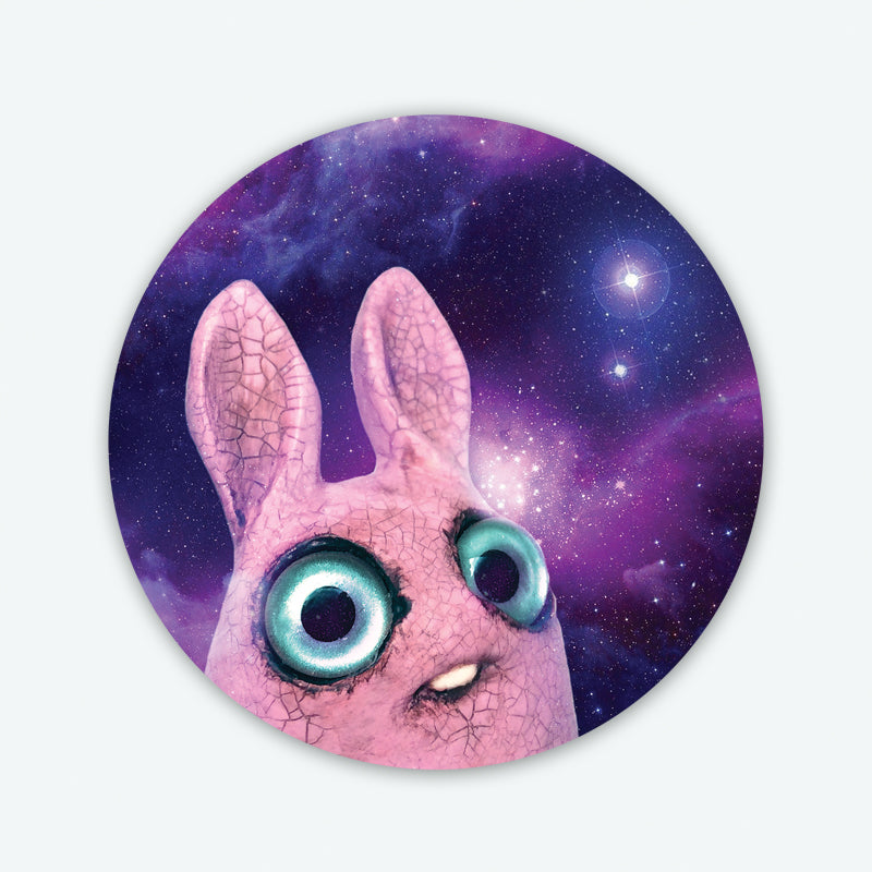 The Pip Galaxy Sticker from Bindlewood Shop features a pink, wide-eyed clay bunny with large blue eyes, gazing upward against a vivid, purple starry galaxy background. This waterproof vinyl sticker is inspired by Amanda Louise Spayd’s creations.