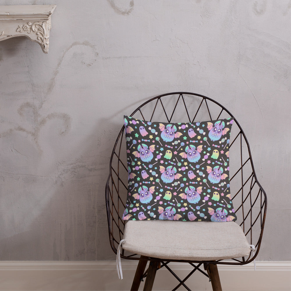 A wire-frame chair with a light-colored seat cushion is placed against a gray wall with subtle designs. On the chair is the Pastel Candy Bat Throw Pillow by Bindlewood Shop, featuring a colorful pattern of cartoon-like animals and symbols on a dark background, adorned with linen-textured pillows for added comfort.
