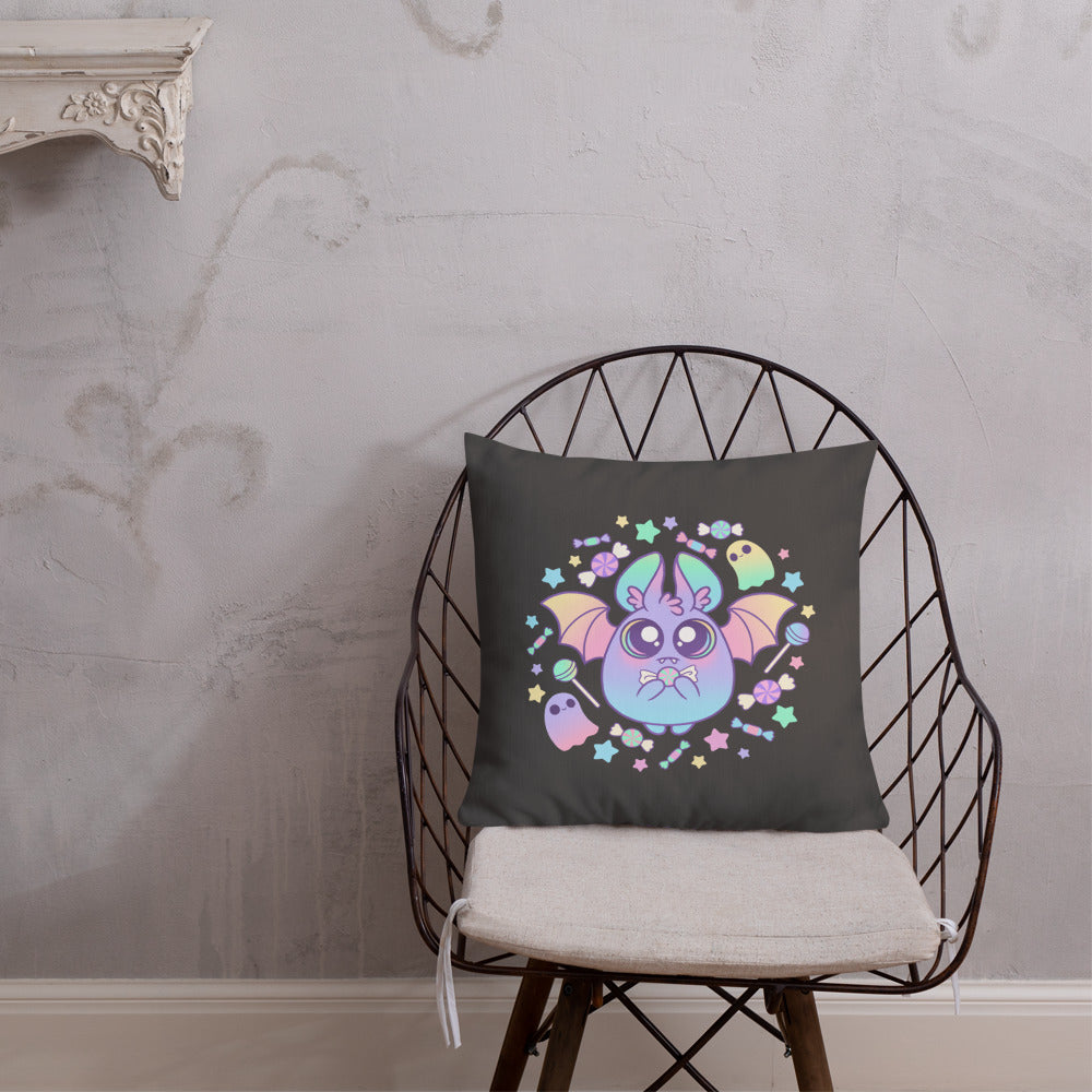 A dark chair with a beige seat cushion holds the Bindlewood Shop's decorative, linen-textured Pastel Candy Bat Throw Pillow, featuring a colorful and whimsical bat with large eyes and wings. The bat is surrounded by stars and two small ghost-like figures. The background of the room is light gray with subtle decorative patterns.