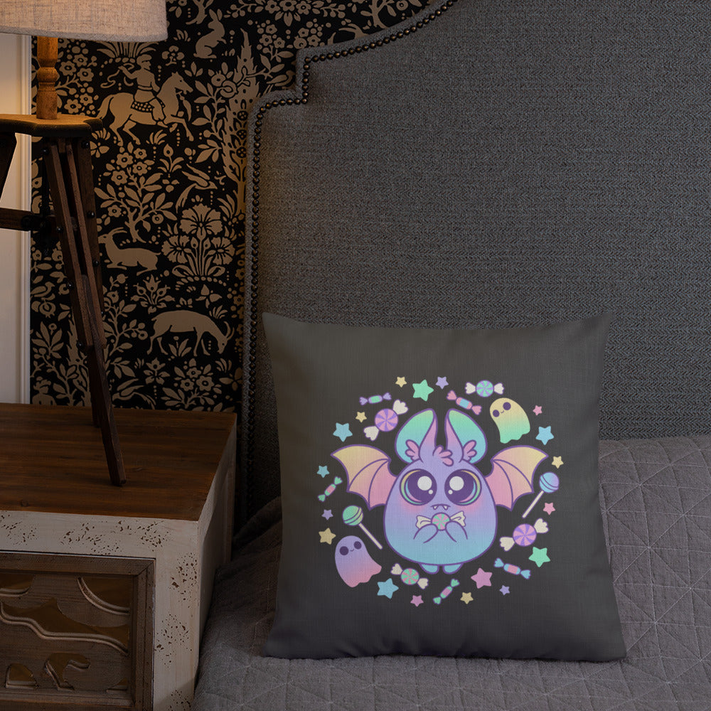 A decorative linen-textured Pastel Candy Bat Throw Pillow from Bindlewood Shop, featuring a cute, colorful central bat with two ghosts surrounded by pastel stars and flowers, rests on a gray bedspread. A nightstand with a wooden lamp and patterned wallpaper with animal motifs are in the background.