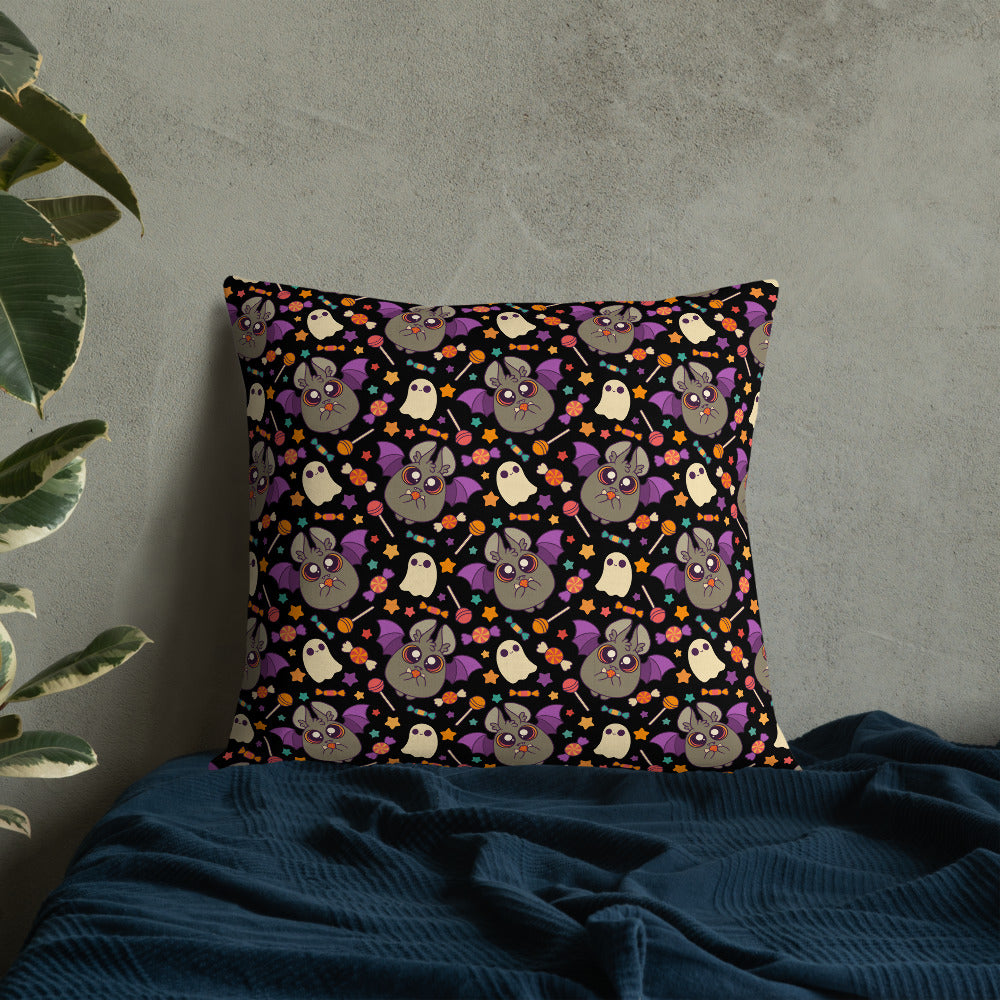 The Candy Bat Throw Pillow by Bindlewood Shop is a square cushion with a black background adorned with a vibrant pattern of cartoon owls and bats in shades of purple, grey, and yellow. Designed by Amanda Louise Spayd, this pillow is set against a concrete wall, resting on textured dark blue fabric with a plant visible on the left.