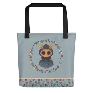 The Twilight Sprite Tote by Bindlewood Shop is a light blue, sturdy tote bag adorned with an illustration of an adorable character with big eyes, dressed in a cloak, and encircled by a floral design. Made from 100% spun polyester fabric, the bottom edge features a floral border on a white background. The handles are black and showcase the Bindlewood logo.