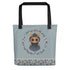 The Twilight Sprite Tote by Bindlewood Shop is a light blue, sturdy tote bag adorned with an illustration of an adorable character with big eyes, dressed in a cloak, and encircled by a floral design. Made from 100% spun polyester fabric, the bottom edge features a floral border on a white background. The handles are black and showcase the Bindlewood logo.