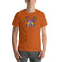A person wearing the soft and lightweight Donut Bat Tee from Bindlewood Shop, which features a cartoon design of a purple bat with large eyes surrounded by colorful candies on an orange background, stands against a plain white backdrop. Crafted for extra comfort, the t-shirt is made from combed and ring-spun cotton.