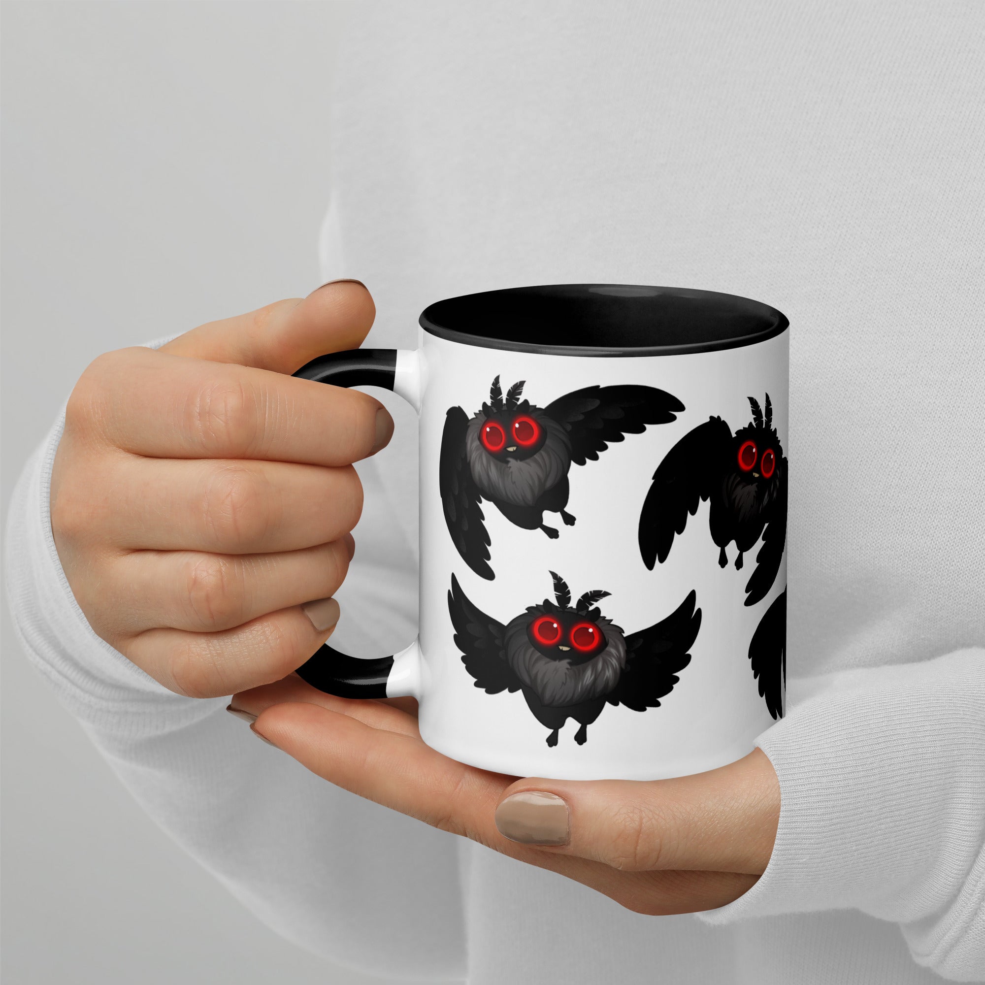 A cryptozoology researcher holds a white Bindlewood Shop Mothman Cryptid Mug with a black interior, featuring cute, black, wide-eyed owl-like creatures with red eyes. The researcher is wearing a white long-sleeved shirt and has neatly manicured nails in a neutral shade. This Mothman Cryptid Mug design evokes the whimsical style of Amanda Louise Spayd.