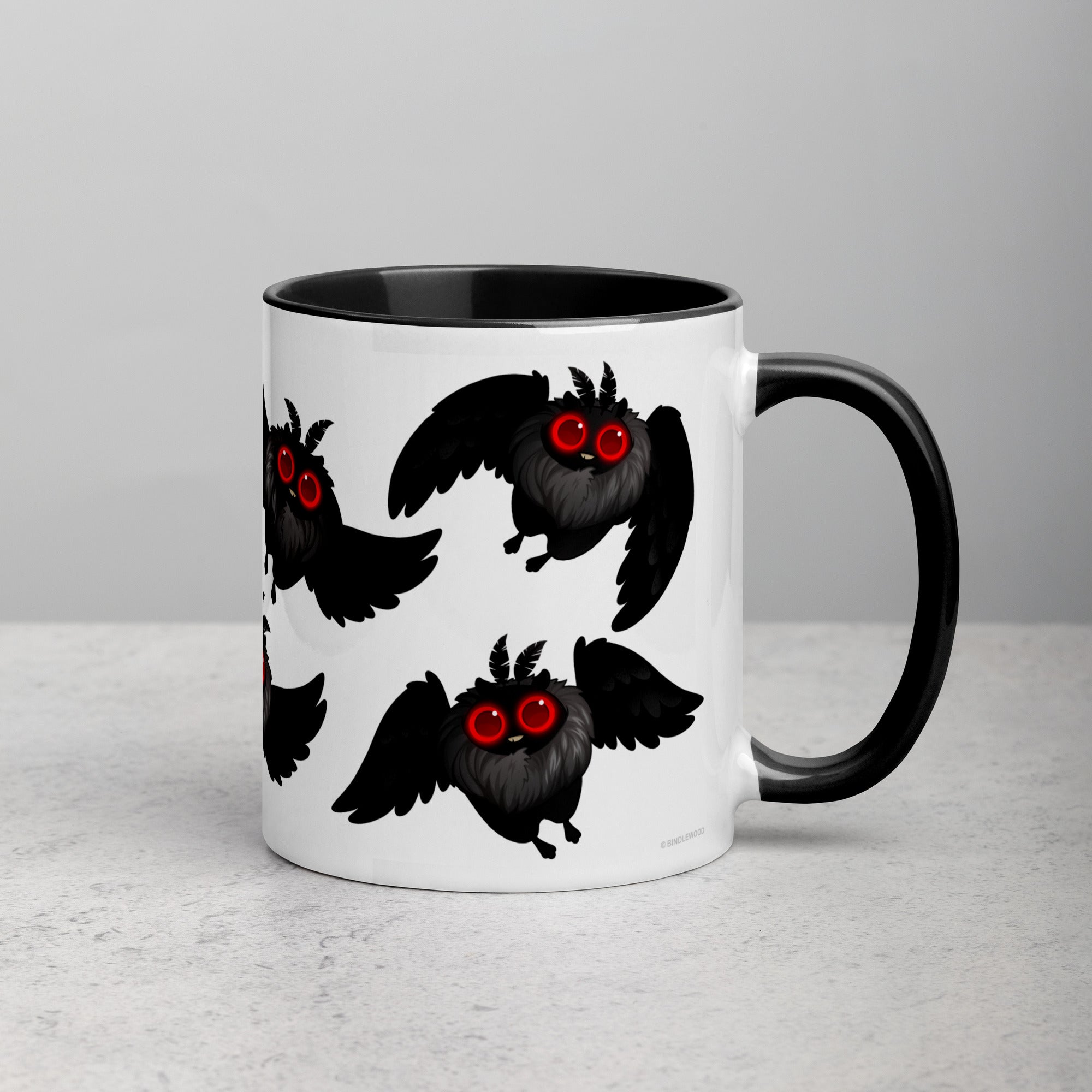 A ceramic Mothman Cryptid Mug from Bindlewood Shop with a black handle and interior, featuring a pattern of cartoonish owl-like creatures with red eyes and outspread wings. The mug is placed on a white surface against a neutral background, making it perfect for any cryptozoology researcher or fan of Amanda Louise Spayd.