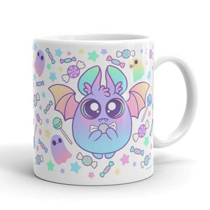 The Pastel Candy Bat Mug from Bindlewood Shop showcases a delightful, round creature with large eyes, bat wings, and holding a bow. The background is beautifully decorated with colorful stars, candies, and small ghost-like figures in soft pastel hues. This whimsical and playful ceramic mug is also dishwasher and microwave safe.